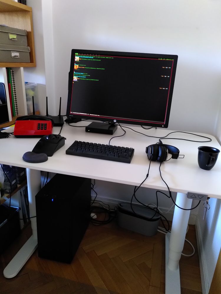 A small white desk with a large black tower computer under it. On
top is, from left, a red touchtone telephone, a wifi AP with antennas
sticking up, a small black computer, a black trackball with a huge
ball, a black keyboard, a black monitor seemingly floating above it
all. On the side there's a pair of black headphones and a black mug.
On the screen is Emacs with a Fedi feed in green on black, but with
some avatar images.