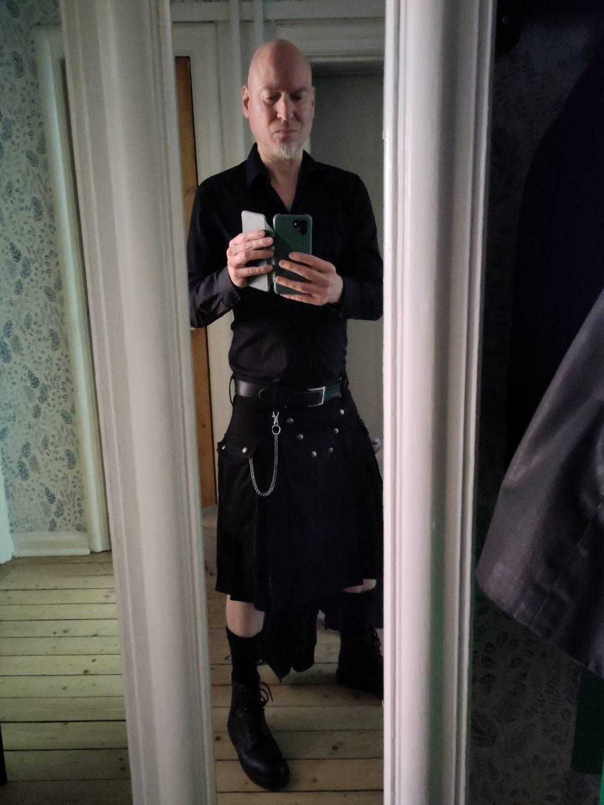 Mirror view of a pale, bald man in a black dress shirt and black
kilt, black socks, black booths, large keychain. He's holding up a
phone and taking this picture.