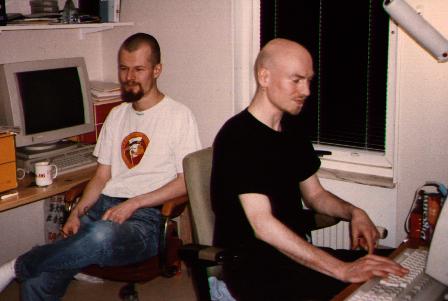 Two pale dudes, one with very short hair and beard in white t-shirt
and blue jeas, one with shaved head and short beard dressed in black,
both in front of screens