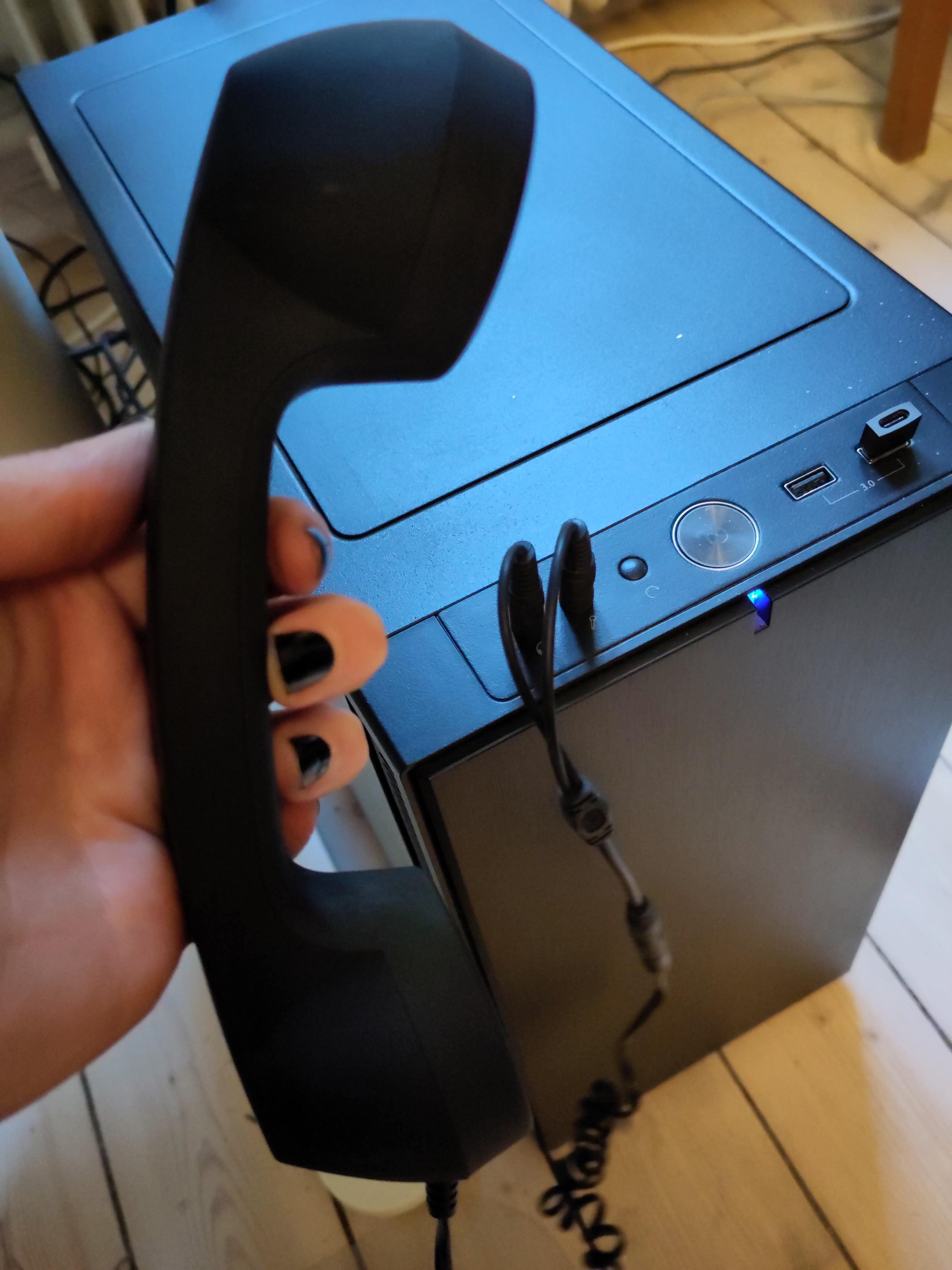 Pale hand with black nailpolish holding a phone handset in front of
a black towercomputer on a wooden floor.